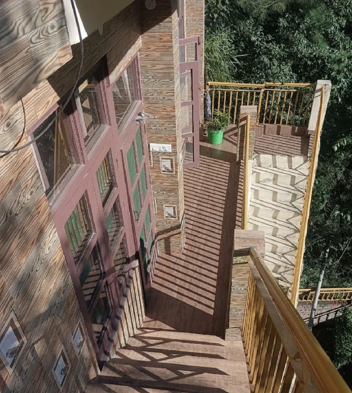 A wooden staircase leads from a doorway, featuring a railing that offers safety. Natural light casts shadows on the steps, highlighting the texture of the walls. Lush greenery is visible in the background, creating a serene environment.