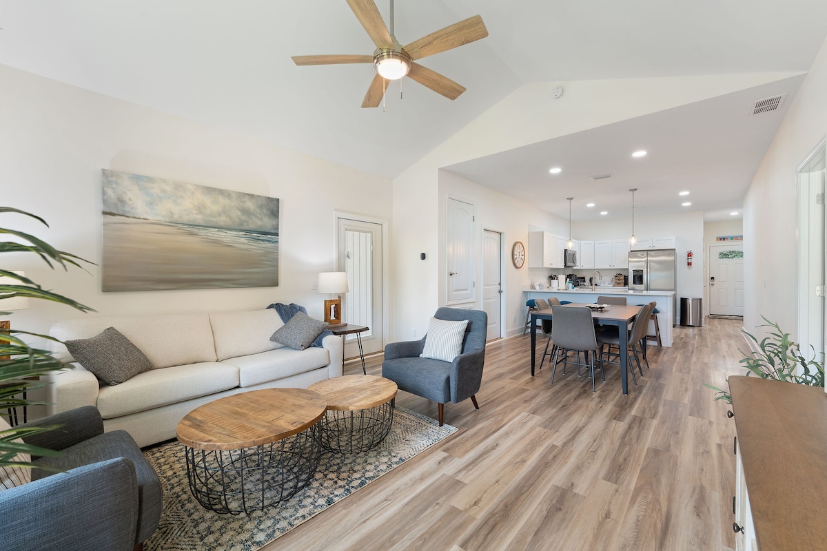 An open-concept living area features a light-colored sofa with two dark armchairs. A wooden coffee table is centered on a patterned area rug. The dining area showcases a modern table with four chairs, adjacent to a fully equipped kitchen with appliances visible in the background.
