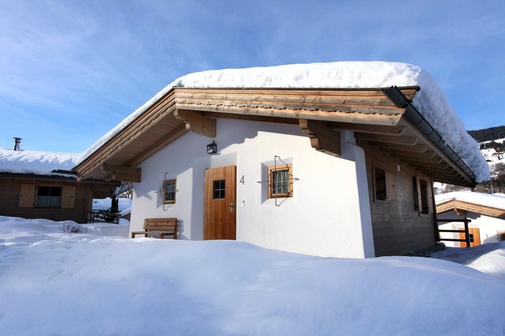 A charming chalet is shown with a sloped roof covered in snow. The building features wooden accents, a front bench, and a wooden door with a small window. Surrounding snowbanks create a serene winter landscape.