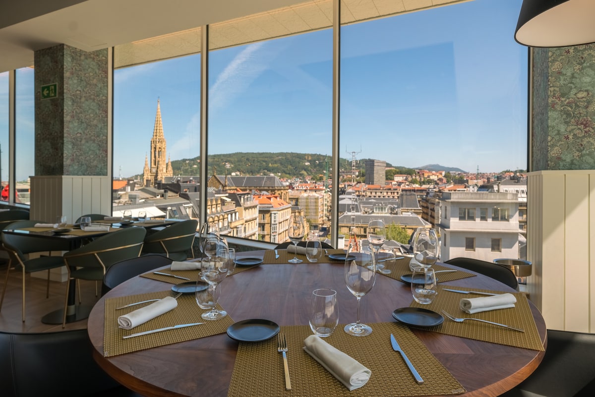 A dining area is presented with a round wooden table set for guests, featuring neatly arranged plates, glassware, and napkins. Large windows showcase panoramic views of the city, with a prominent church steeple in the distance and a clear blue sky overhead.