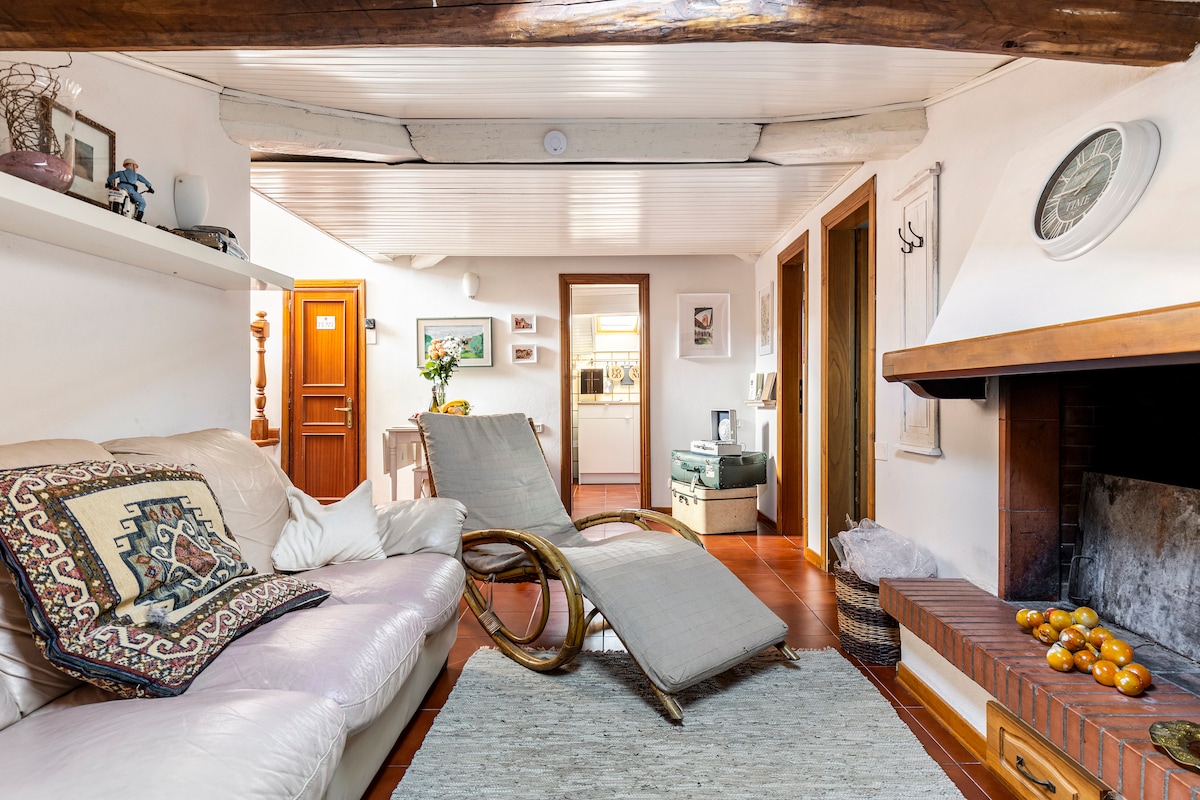 A cozy living area features a soft leather sofa and a stylish lounge chair, inviting relaxation. A rustic fireplace with a decorative mantle is positioned nearby. The space is adorned with subtle art pieces and shelves, contributing to a warm and homey feel.