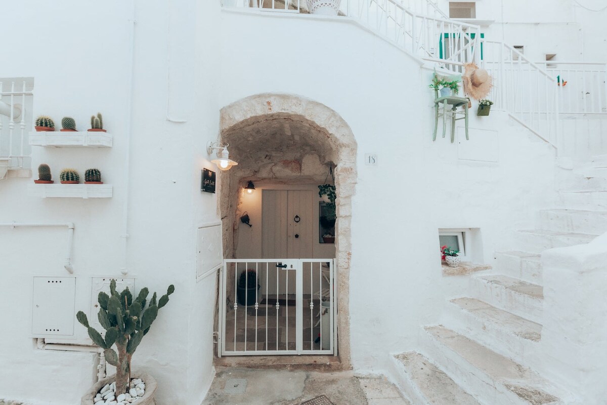 A charming white exterior features an arched entrance framed by a decorative railing. Potted plants adorn the walls, and a staircase leads to an upper area. The entrance door is inviting, with a small decorative wreath visible.