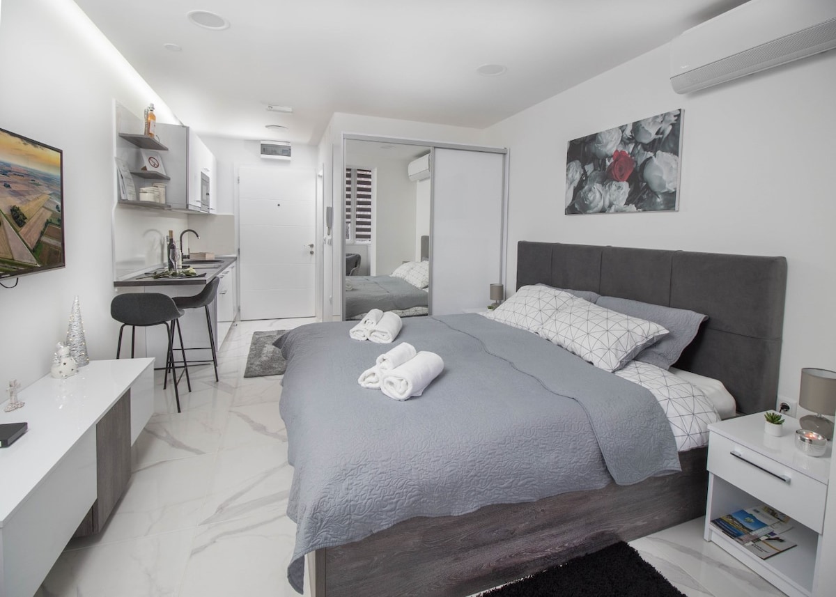 A modern studio apartment features a spacious bed with a gray coverlet and neatly arranged towels. A kitchenette is visible to one side, alongside bar stools against a small counter. A mirrored wardrobe reflects the room, and the décor includes floral art and soft lighting.