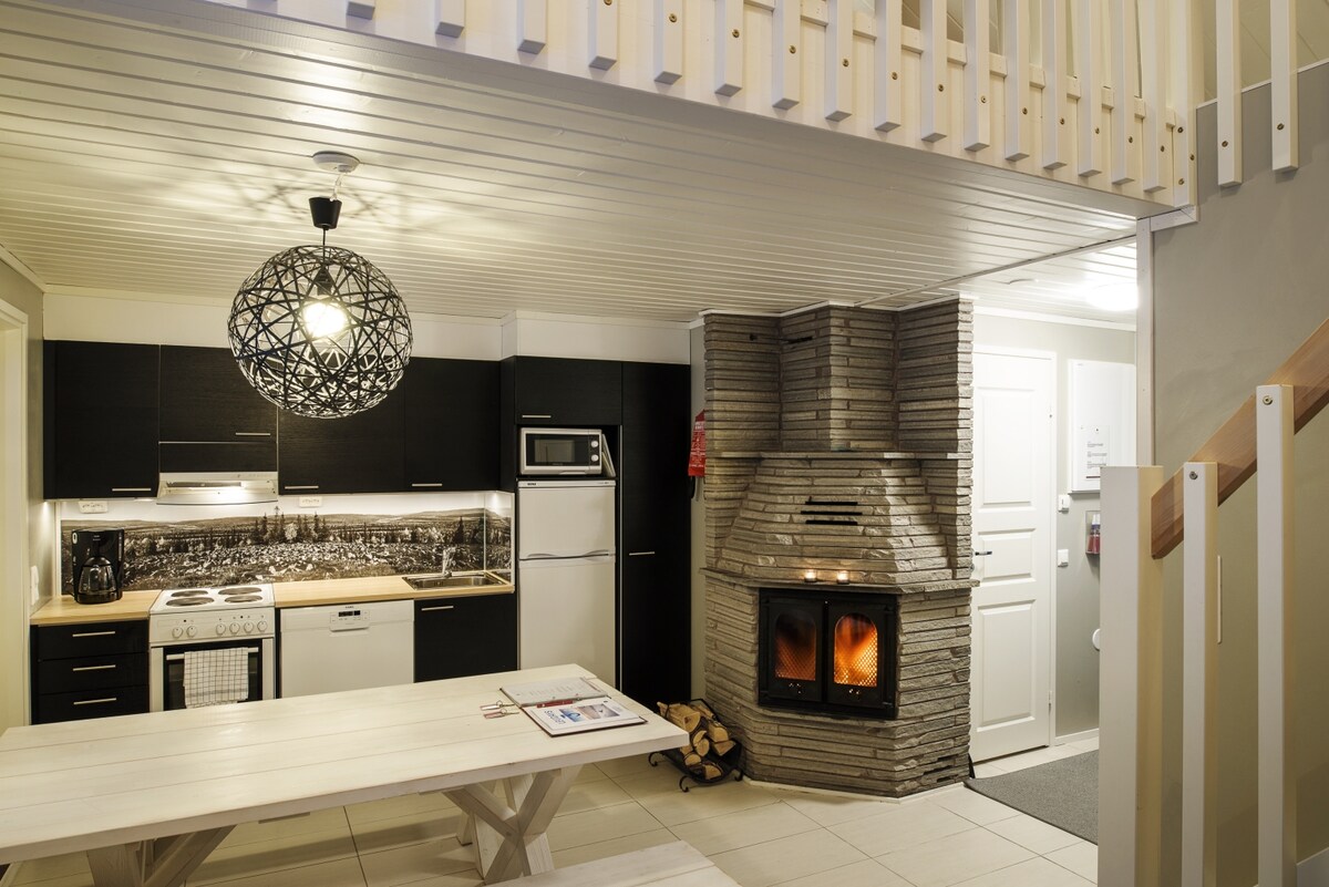 A modern kitchen area is seen, equipped with a stove, refrigerator, and ample cabinetry. A dining table made of light wood is positioned in the center, and a cozy stone fireplace is featured in the corner, with logs neatly stacked beside it. A lofted sleeping area can be glimpsed above.