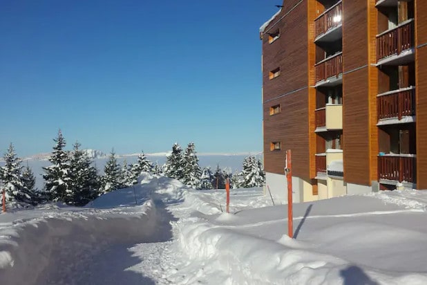 A snowy landscape surrounds a multi-story building with wooden accents. The path through the snow leads towards the entrance, where balconies can be seen on the upper floors. Spruce trees provide a natural backdrop against the clear blue sky.