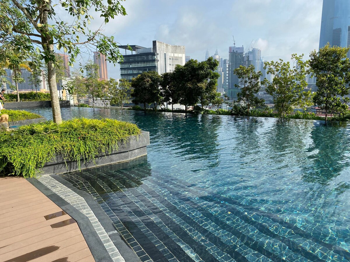 An infinity lap pool is seen with clear water reflecting the surrounding greenery and skyline. The pool features a clean edge that appears to blend seamlessly into the horizon. Lush plants and trees line the area, providing a serene ambiance.