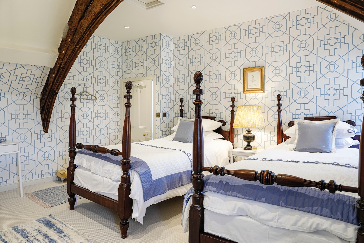 The twin guest room is characterized by two wooden four-poster beds arranged symmetrically. A patterned wallpaper in blue and white adds visual interest, while a lamp casts soft light on a side table. Exposed wooden beams create an inviting architectural detail above.