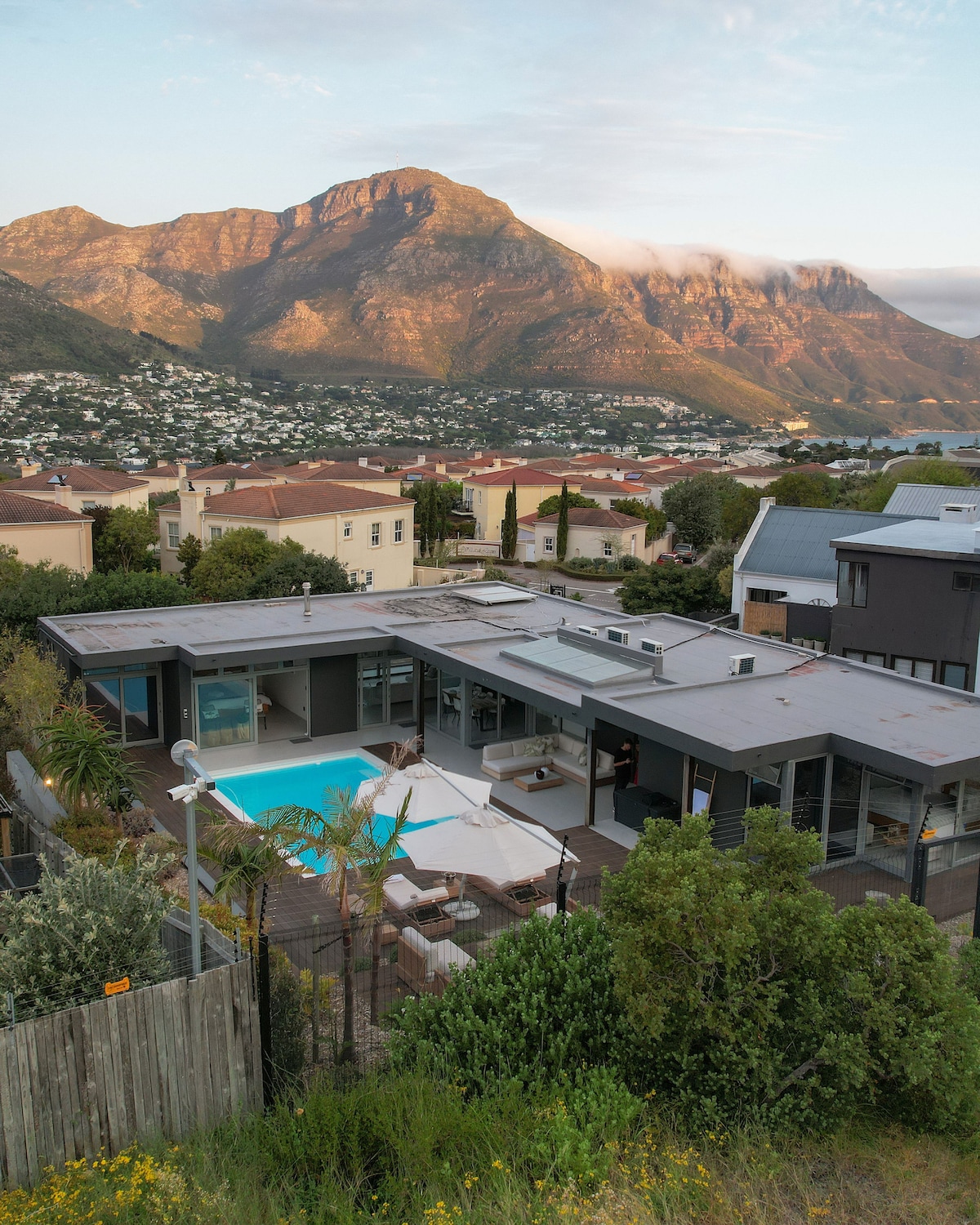 The modern Bauhaus-style house is set against a backdrop of stunning mountains. A well-maintained pool is visible in the outdoor area, surrounded by seating arrangements. Lush greenery and a secure fence enhance the outdoor space, creating a tranquil retreat in a scenic neighborhood.