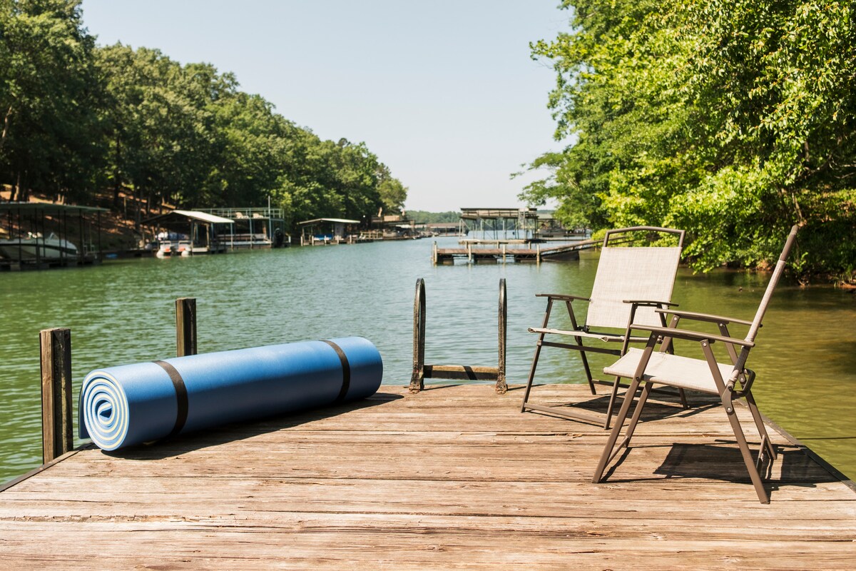 A wooden dock extends over calm waters, featuring a blue flotation mat rolled out on the surface. A pair of light-colored folding chairs are positioned nearby, offering a place to relax while enjoying views of the water and surrounding greenery.