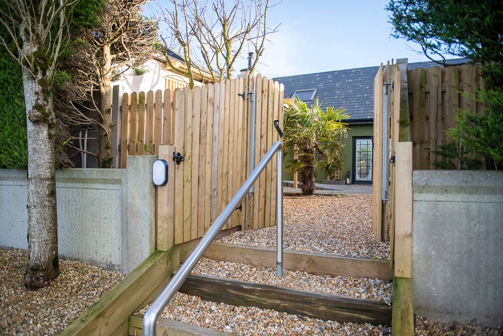 A wooden gate opens to a gravel pathway leading into a well-maintained garden area. Steps are flanked by low stone walls, with lush greenery visible on either side. A glimpse of the home is visible in the background, showcasing architectural design.