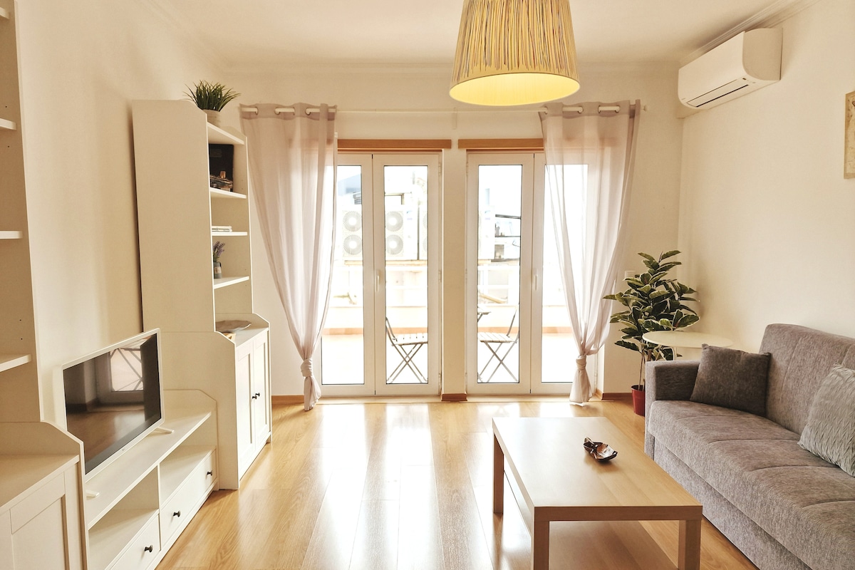 The living room features a comfortable sofa bed, a coffee table, and a television unit against a wall. Two large windows with light drapes allow natural light to fill the space, providing views of the terrace and greenery. A decorative plant adds a touch of warmth.