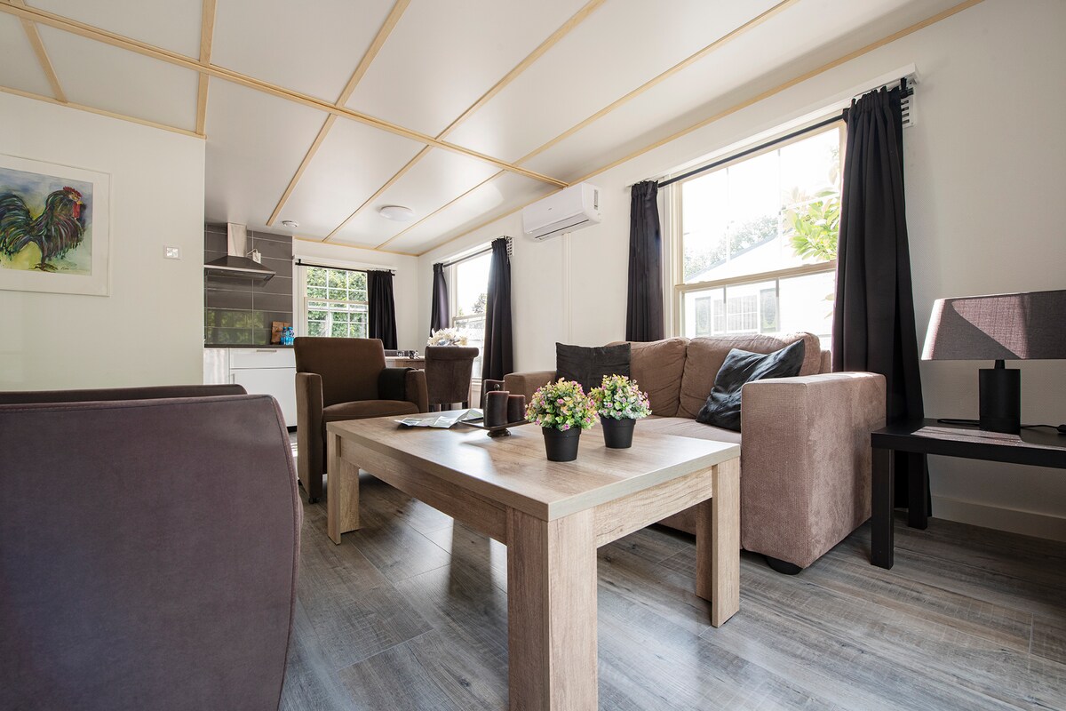 A spacious living area is showcased with comfortable seating on brown couches and matching armchairs. A wooden coffee table sits at the center, adorned with potted plants. Large windows invite natural light, complemented by dark curtains, enhancing the warm and relaxing atmosphere.
