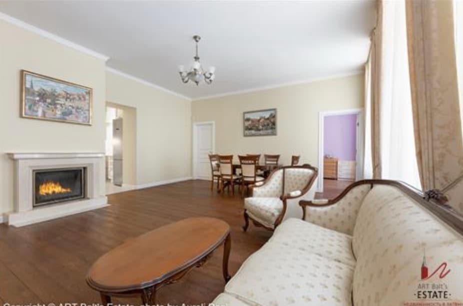 A spacious living area features a decorative fireplace and elegant furnishings. A comfortable sofa and a wooden coffee table are arranged in front of the fireplace. In the background, a dining table with chairs is visible, alongside a soft-colored wall with artwork.