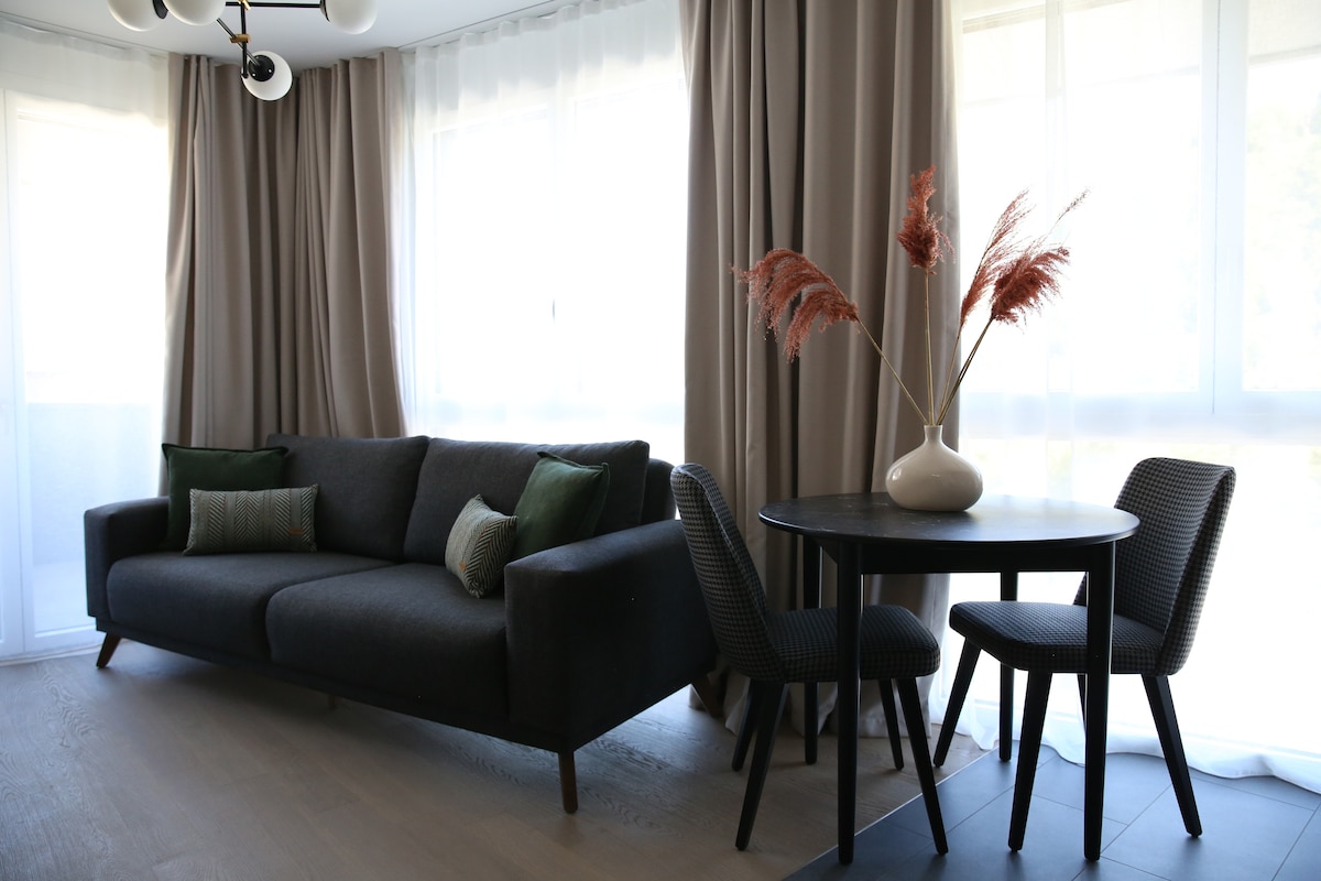 A modern sitting area features a dark sofa adorned with green cushions, complemented by a round dining table and two chairs. Natural light filters through large windows, enhancing the neutral-toned curtains and creating a comfortable ambiance.