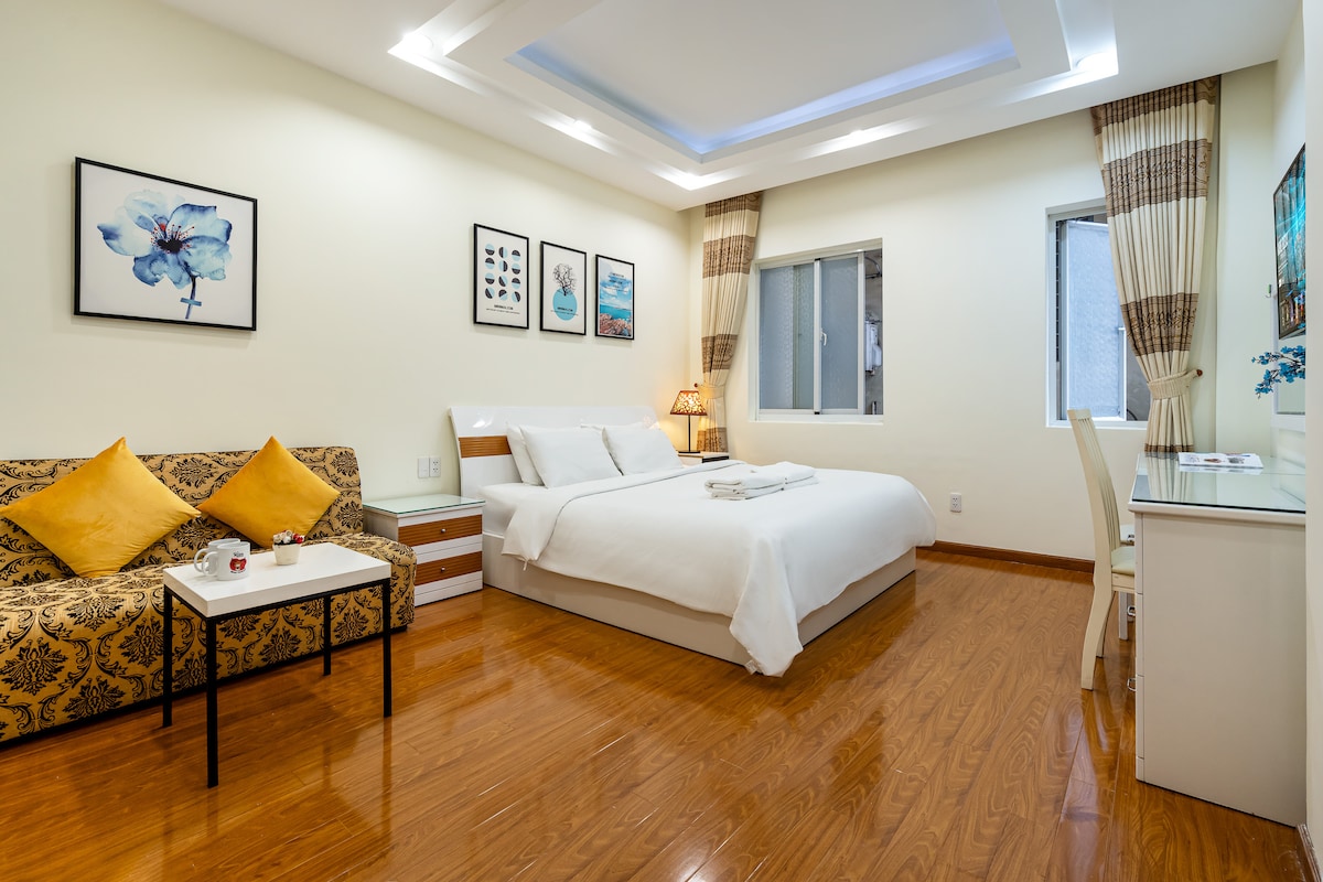 A bright and airy room features a spacious bed dressed in crisp white linens, accompanied by decorative pillows. A patterned sofa sits beside a small table, and artwork is displayed on the walls. Natural light enters through a window, complemented by light-colored curtains.