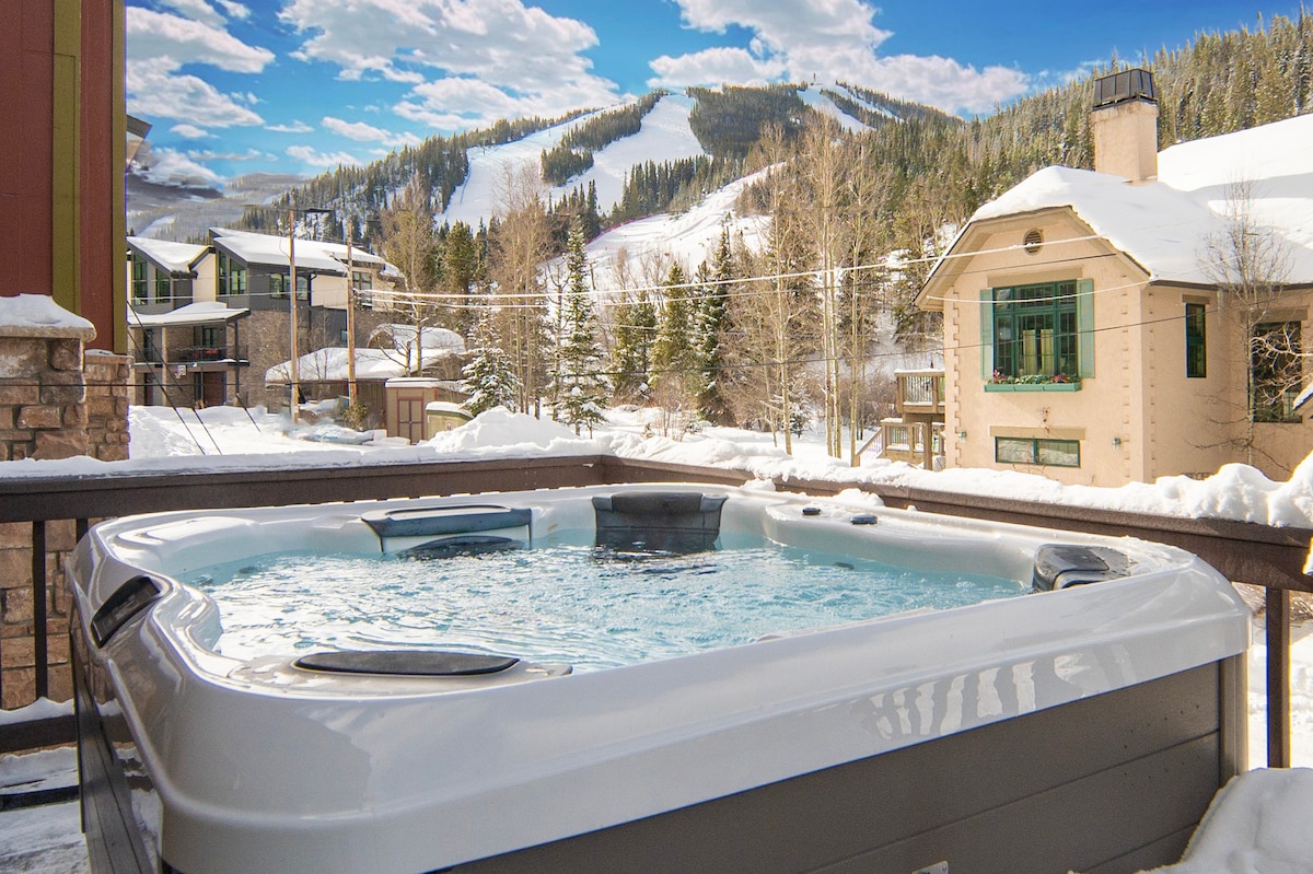 A hot tub is positioned on a deck, providing views of snow-covered mountains and trees in the background. The scene captures a serene winter landscape with scattered clouds in the sky, suggesting a relaxing outdoor experience.