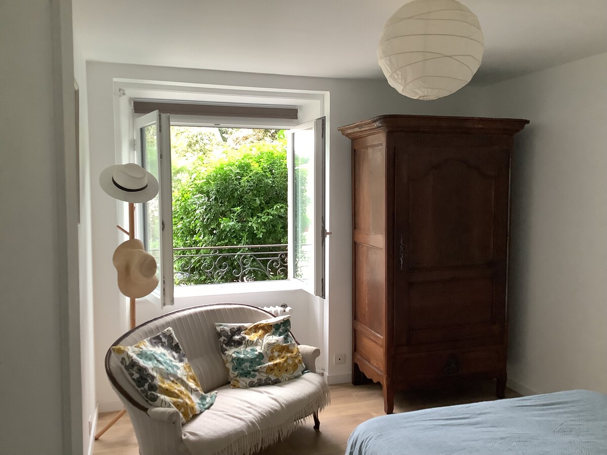 A charming room features a cozy seating area near a window with doors that open to a greenery view. A vintage wardrobe stands against the wall, and decorative pillows add a touch of color to the seating. A soft light fixture hangs from the ceiling.