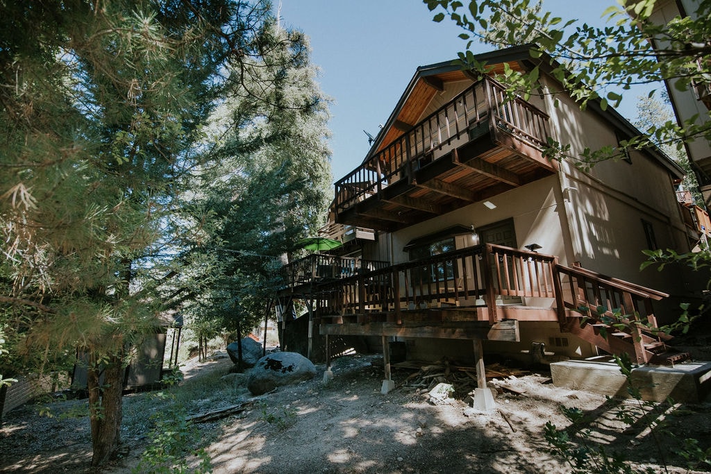 The exterior of the cabin features a two-story structure surrounded by trees, showcasing two balconies with wooden railings. A gravel area is visible below, with natural stones and vegetation, creating a peaceful outdoor setting.