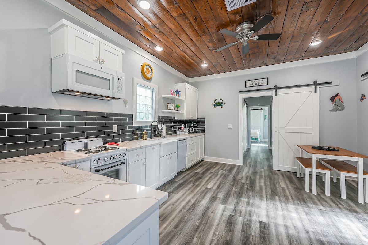An open concept kitchen and living area is highlighted by modern appliances and a spacious countertop. A wooden table with seating for two is positioned against the wall. Natural light enters through a window, and the ceiling features exposed wood beams.