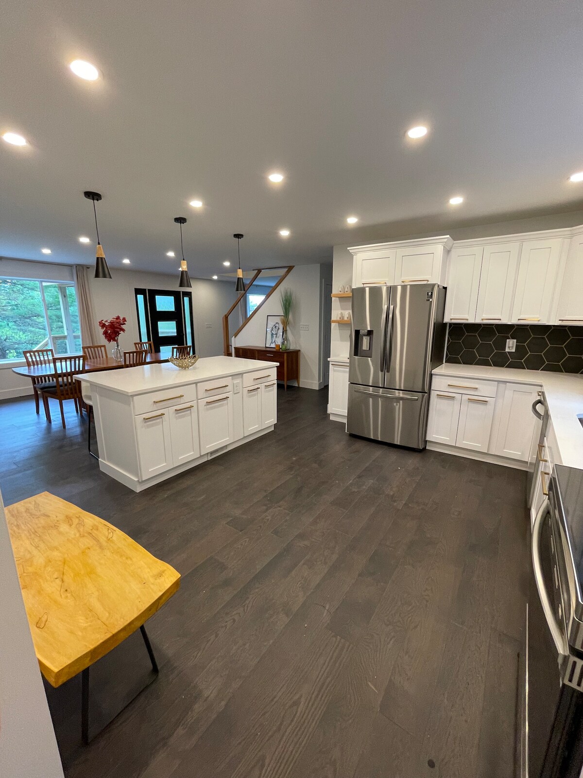 A modern kitchen features white cabinetry, a large island with seating, and stainless steel appliances. Ample lighting enhances the space, while a staircase leads to the upper level in the background. A dining area with wooden chairs is visible near a large window, inviting natural light.