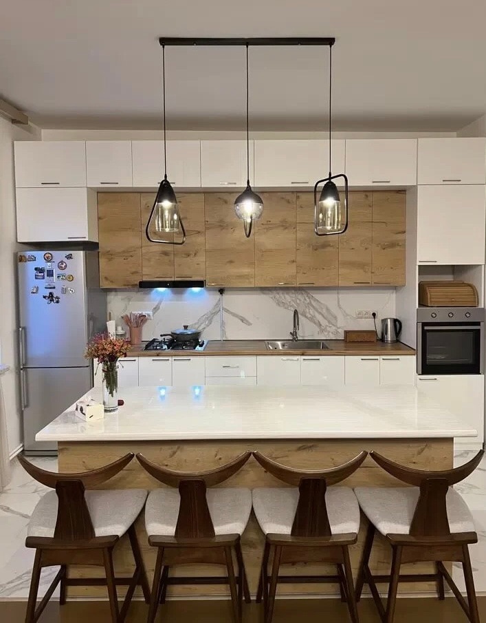 A modern kitchen is highlighted by a large island with seating for four. Elegant pendant lights hang above the island, illuminating the space. White cabinetry contrasts with warm wood finishes, while stainless steel appliances and marble countertops add functionality and style.