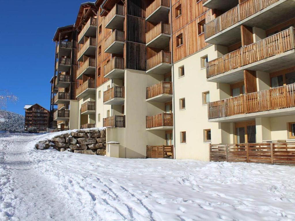 A multi-story building is depicted, featuring a combination of wooden balconies and stone elements. Snow covers the ground, reflecting a clear blue sky. The structure is designed with a rustic alpine style, prominently showcasing several balconies that extend outward.