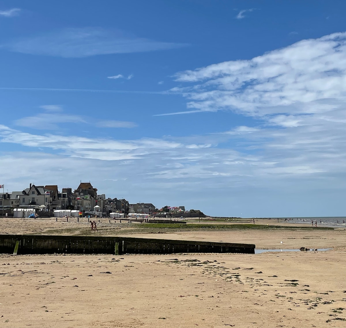 A sandy beach stretches into the distance, framed by a clear blue sky dotted with soft clouds. The shoreline is lined with various buildings, showcasing a mix of architecture. People can be seen enjoying the beach, with a wooden structure extending into the water.