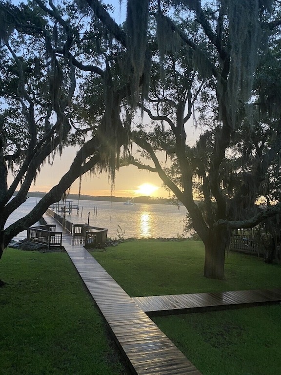 Sunset hues illuminate the waterway, viewed through the branches of mature trees adorned with Spanish moss. A wooden walkway extends towards a 300-foot dock, surrounded by lush green grass and cozy seating areas, creating a serene atmosphere for evening relaxation.