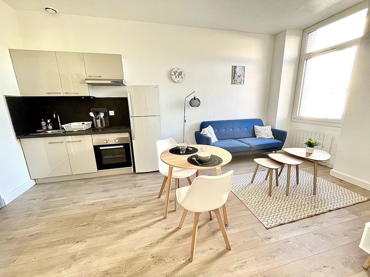 An open living space features a modern kitchen area with essential appliances, including a refrigerator and a stove. A blue sofa offers seating, complemented by a round dining table surrounded by four chairs. Natural light enters through large windows, creating a bright environment.