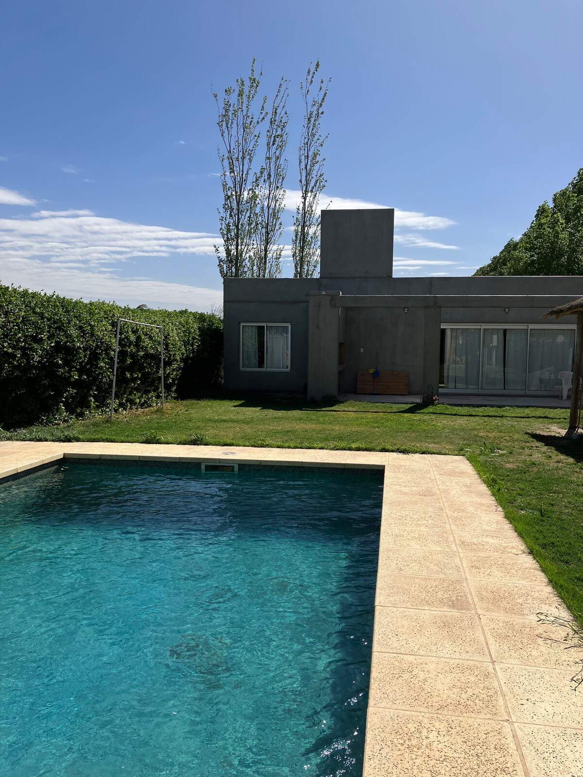A serene outdoor space features a turquoise swimming pool surrounded by a light-colored patio. A modern structure with large glass doors is set against a backdrop of green hedges and tall trees, enhancing the tranquil ambiance.
