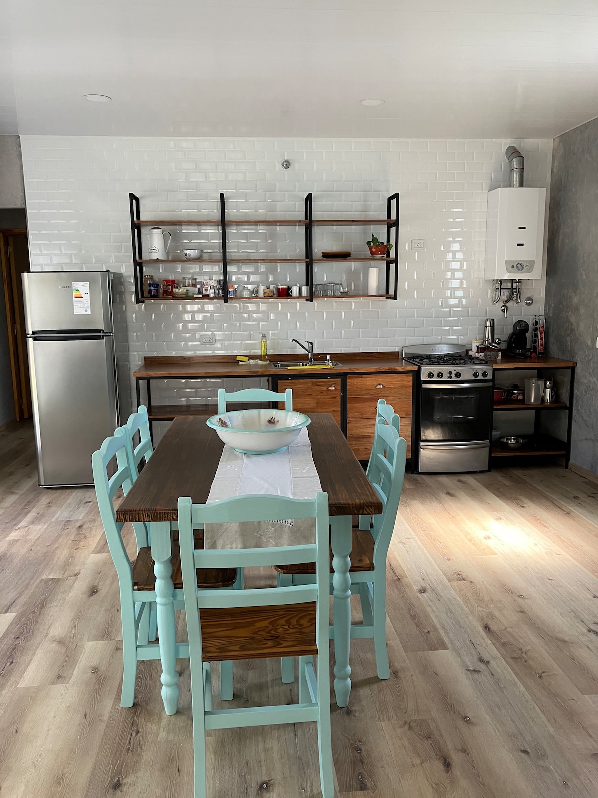 A modern kitchen is illuminated by natural light, featuring a wooden dining table surrounded by light blue chairs. An equipped stove and refrigerator are present, along with open shelving displaying various kitchen items. Wood and tile flooring contribute to the clean, contemporary look.