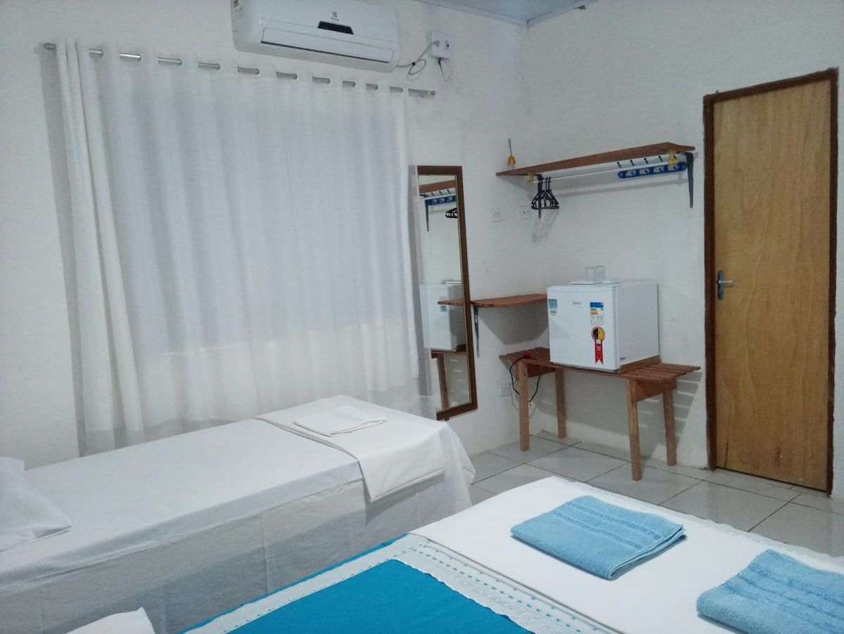 A well-lit room is furnished with two beds dressed in light bedding, featuring blue accents. A small refrigerator is positioned beside a wooden shelf. Natural light filters through a sheer white curtain, enhancing the neutral wall colors and complementing the wood tones of the furniture.