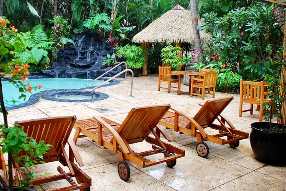 A tranquil outdoor area is shown, featuring a pool surrounded by lush tropical foliage. Four wooden lounge chairs are positioned near the pool, offering a relaxing space. A thatched umbrella provides shade, while a small table and chairs sit nearby, creating an inviting atmosphere.