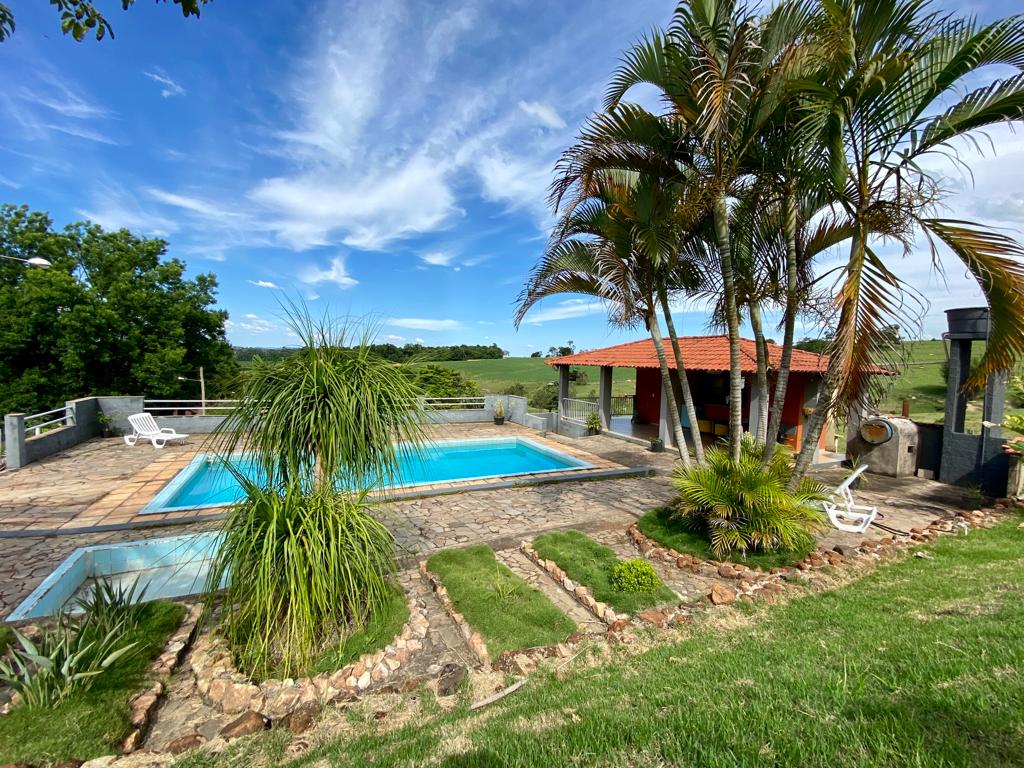 A refreshing swimming pool is surrounded by lush greenery and palm trees, with a stone patio featuring lounge chairs. A shaded area with a red-roofed structure provides a backdrop, and expansive views of the landscape are visible in the distance.