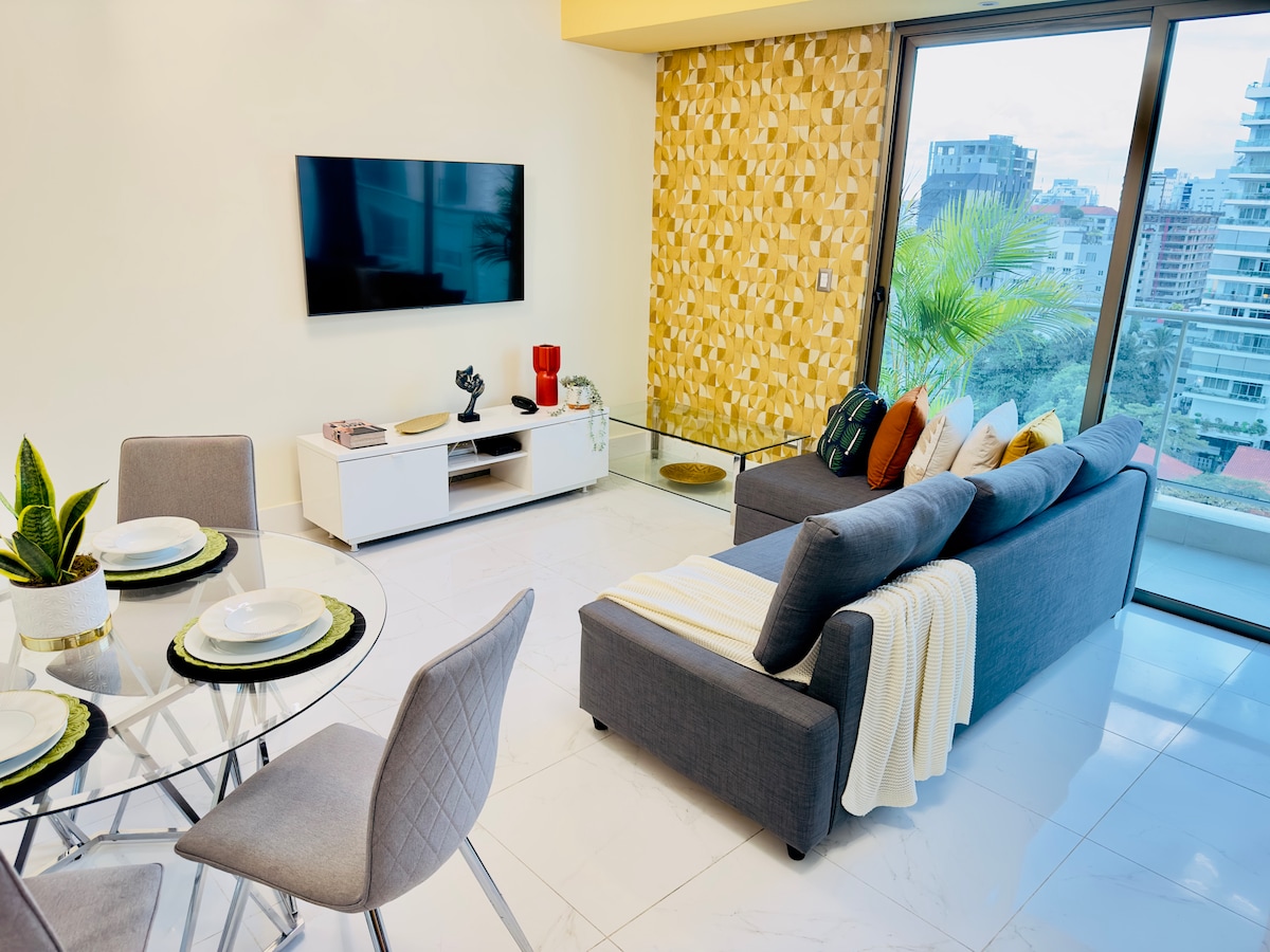 A modern living area features a cozy sectional sofa adorned with a soft throw. A glass dining table is set for four, with stylish place settings. A wall-mounted television is positioned above a minimalist TV console, while large windows provide views of the cityscape.