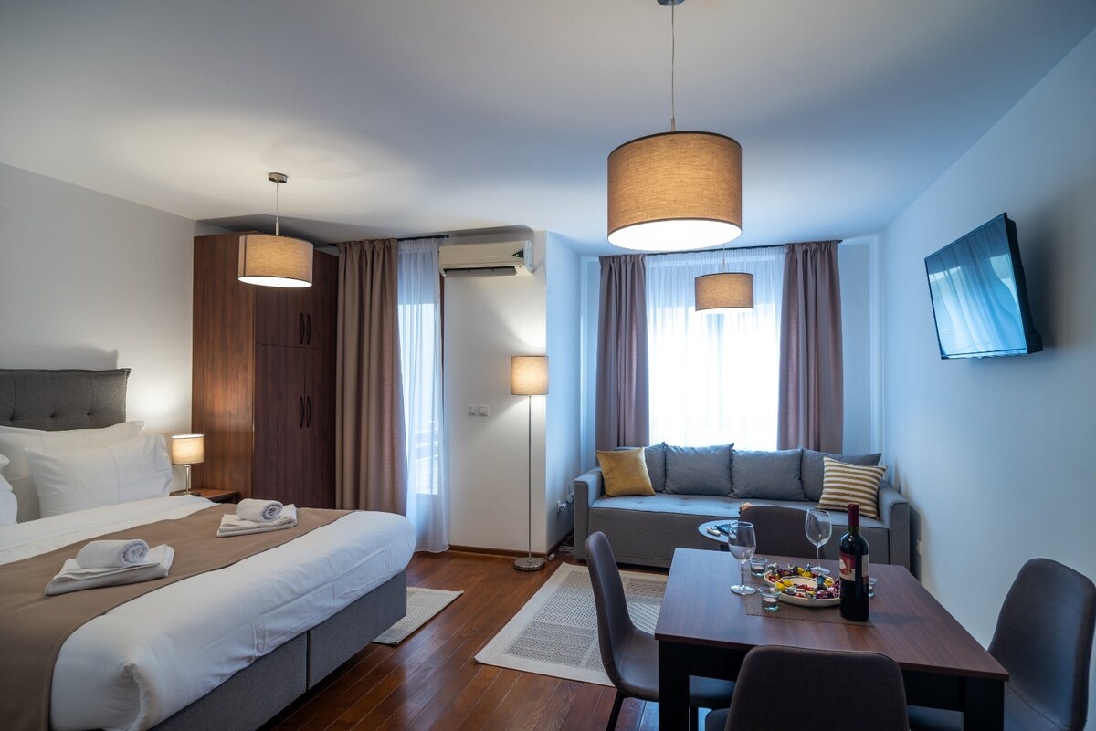 A well-lit studio apartment features a spacious bed with neatly arranged towels. An intimate dining area with a wooden table and chairs is situated next to a cozy seating area. Natural light filters through the windows, complemented by modern light fixtures and soft curtains.