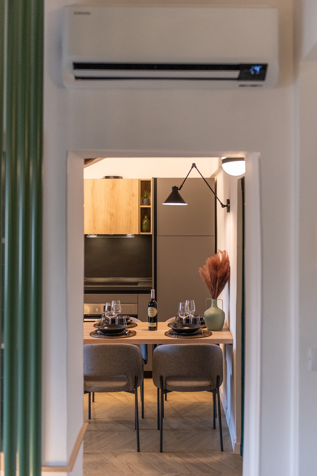 A modern kitchen area is visible through an inviting corridor. A wooden dining table set for two is adorned with wine glasses and decorative elements. Plants and warm lighting complement the contemporary kitchen cabinetry and appliances.