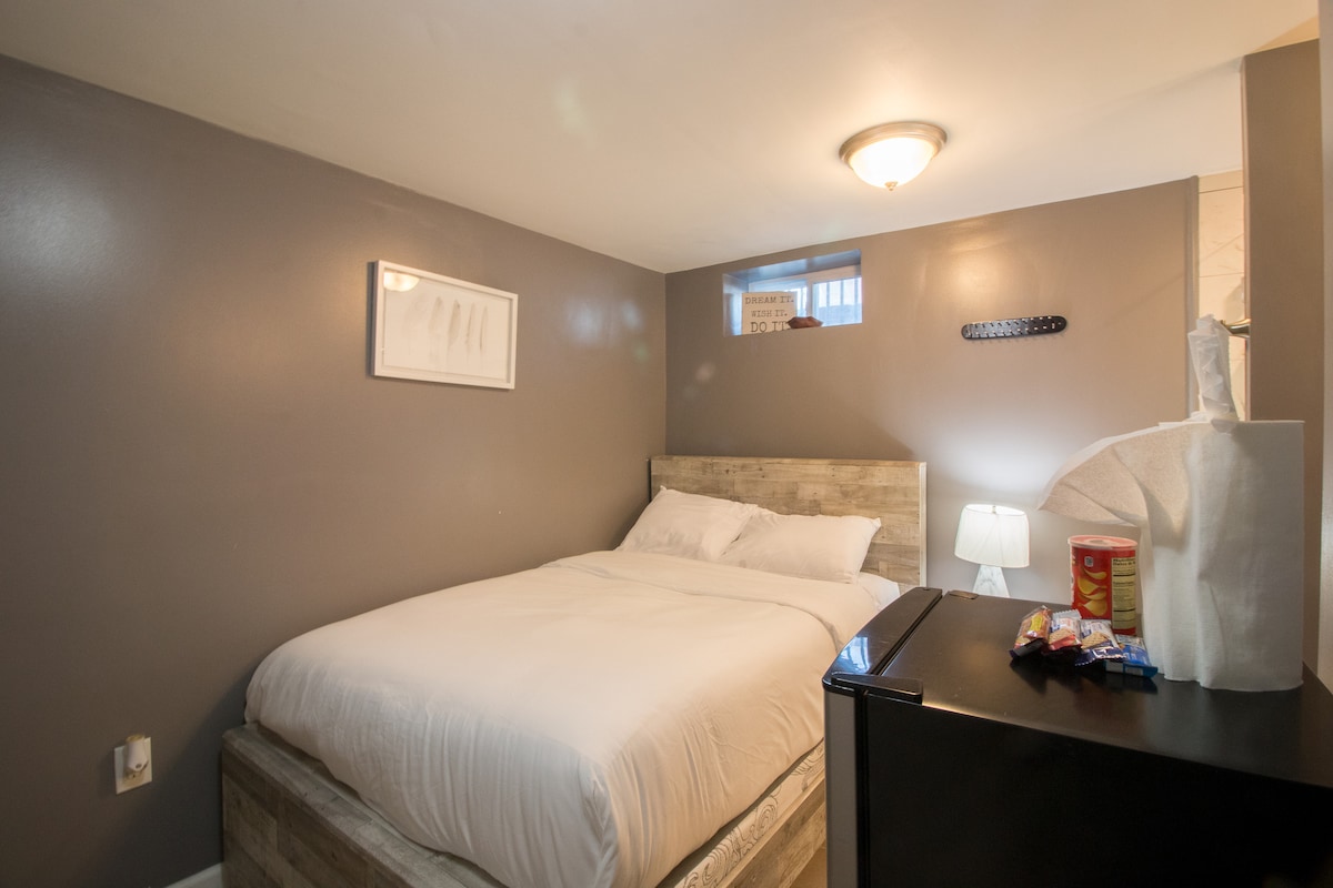 A cozy sleeping area features a full-size bed with white bedding, complemented by a nightstand with a lamp. A small fridge is present beside the bed, while a framed artwork adorns the gray wall. Natural light filters through a small window, enhancing the calm environment.