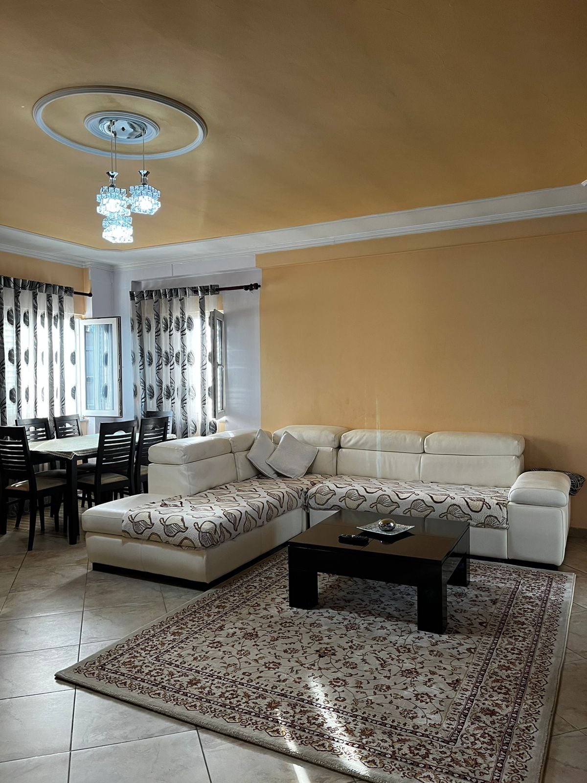 A light-filled living area features a cream-colored sectional sofa with decorative cushions, positioned near a dark coffee table. A patterned area rug lies on tiled flooring. Dining chairs surround a table in the background, with patterned curtains framing large windows.