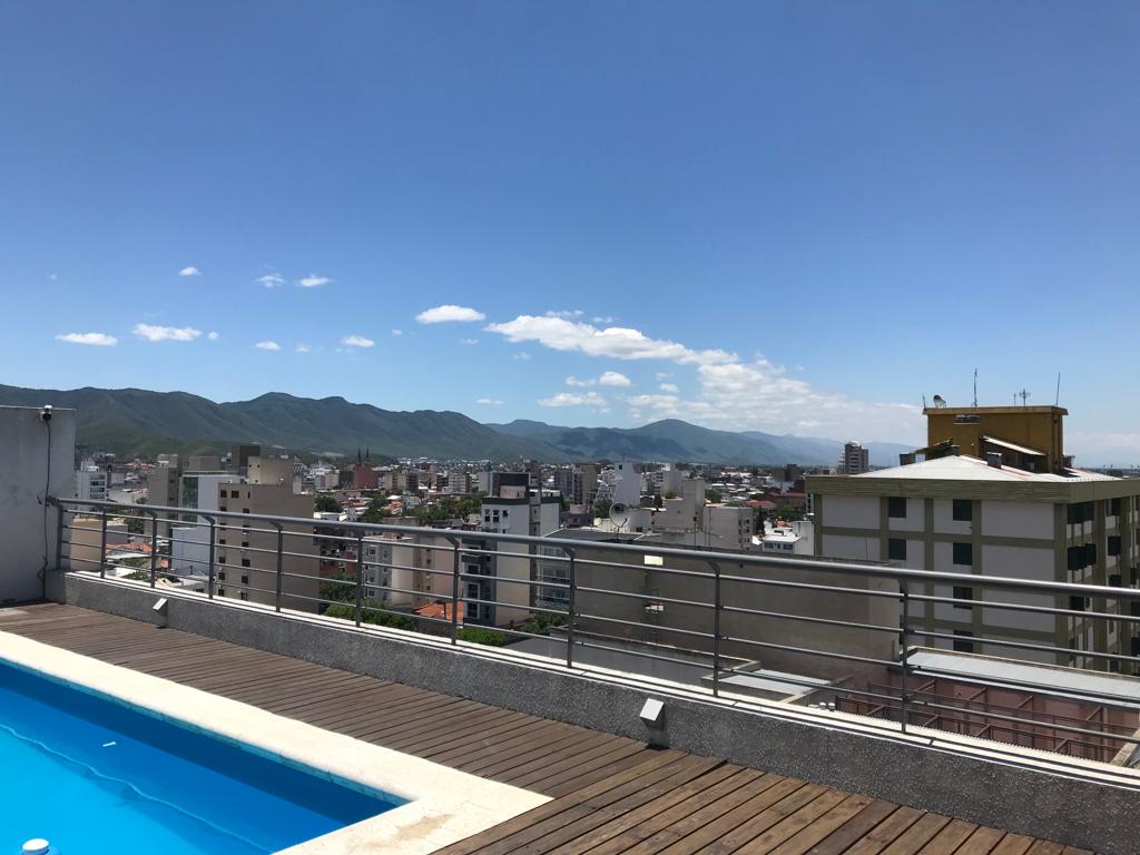 A rooftop area features a rectangular pool bordered by wooden decking. The city skyline stretches out in the distance, complemented by mountains under a clear blue sky with scattered clouds.
