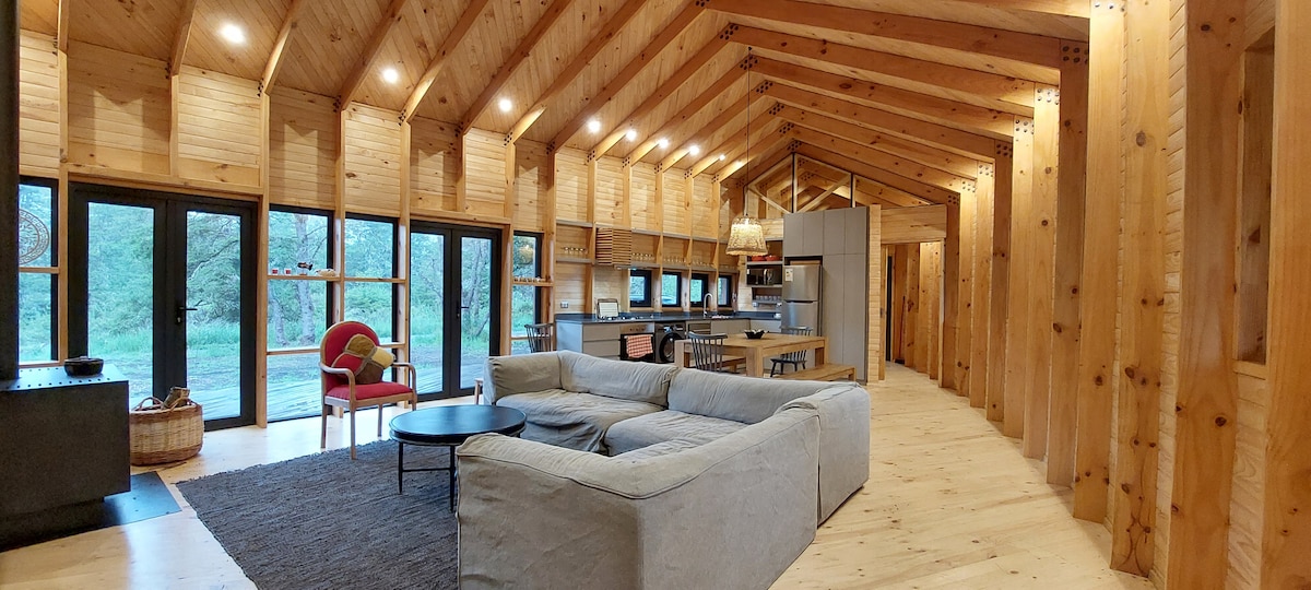 The spacious living and dining area features wooden walls and vaulted ceilings. Large glass doors offer views of the surrounding nature. A comfortable sectional sofa faces a central coffee table, while a well-equipped kitchen is visible in the background, illuminated by soft overhead lights.