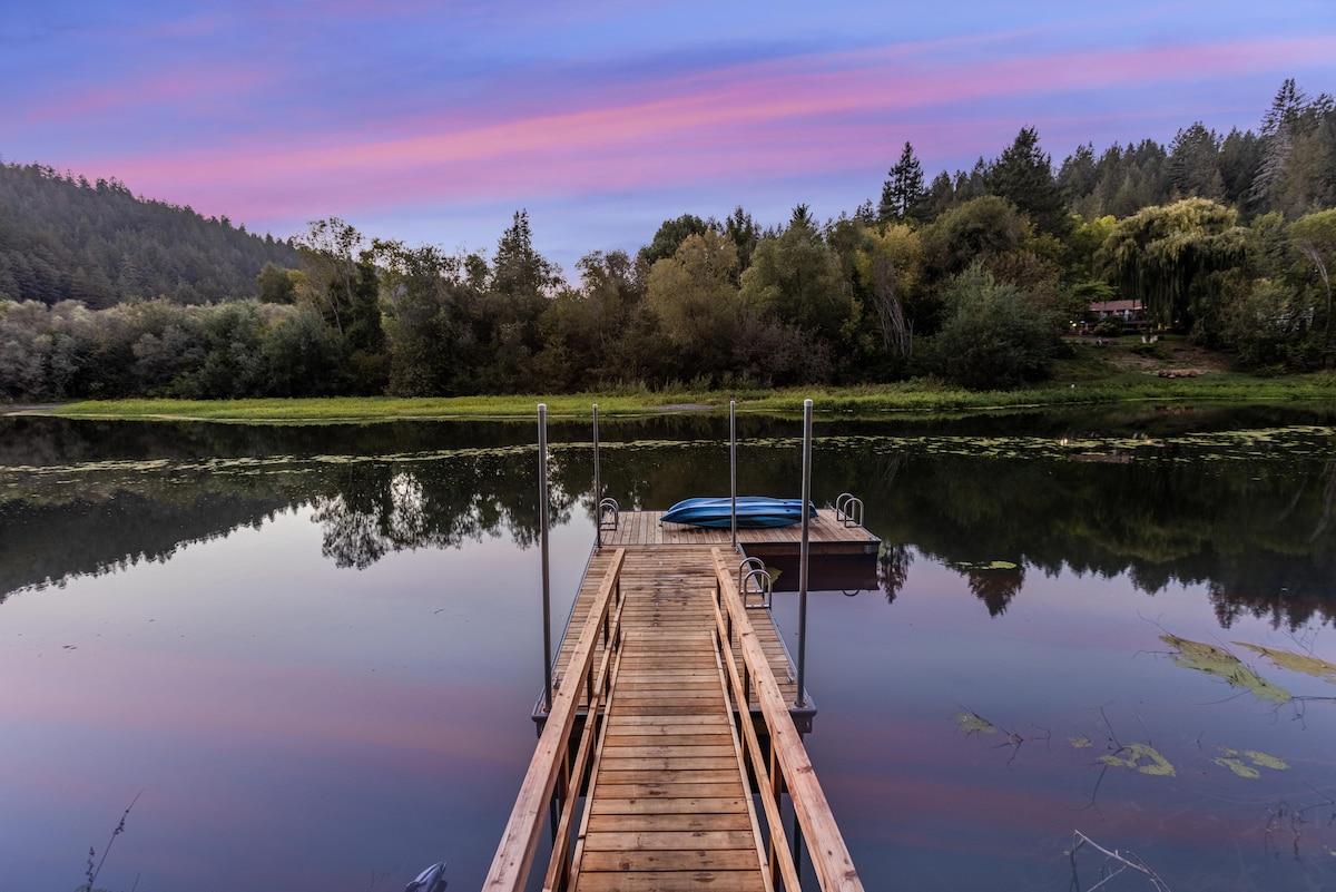 A wooden dock extends into calm water, reflecting the colors of a sunset sky. Lush greenery surrounds the area, with trees lining the far shore. A kayak is visible on the water, adding to the serene natural setting.