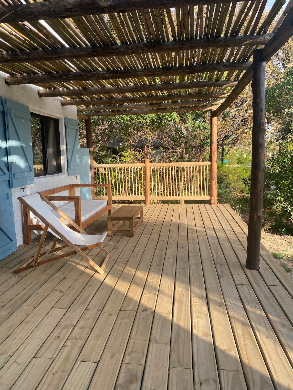 A spacious wooden deck is featured, providing a relaxing outdoor space. Two lounge chairs and a small table are arranged alongside a bench, surrounded by lush greenery. Natural light filters through the open beams of a thatched roof above.