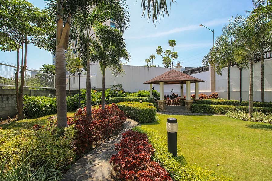 A well-maintained garden area is surrounded by lush greenery and colorful flower beds. A gazebo with a thatched roof provides a shaded spot for relaxation, while a stone pathway leads through the vibrant landscape. Tall palm trees and neatly trimmed bushes enhance the serene ambiance.