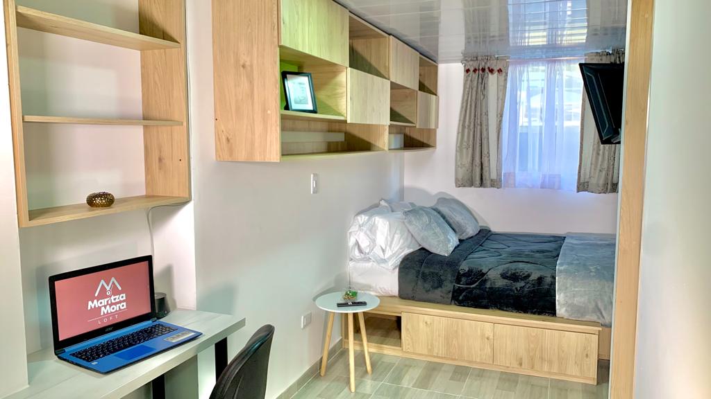 A well-organized room features a spacious bed with plush bedding, complemented by modern wall shelves. A compact workspace with a desk and chair is positioned nearby, alongside a small side table. Natural light enters through a window adorned with sheer curtains.
