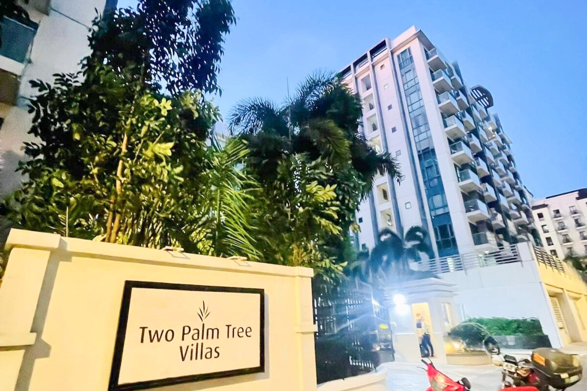 The entrance to Two Palm Tree Villas is showcased, featuring a prominent sign against a backdrop of lush greenery. A modern building with multiple balconies stands tall, illuminated by exterior lighting as dusk settles in, creating a serene atmosphere.