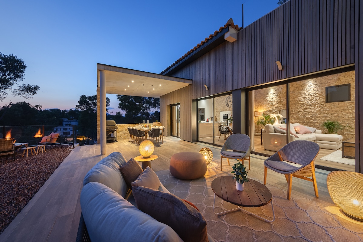 An outdoor seating area features a circular table surrounded by cushioned chairs, complemented by a soft ottoman. Through large glass doors, a well-lit living space is visible, showcasing natural materials and a warm ambiance. Soft lighting enhances the serene evening atmosphere.