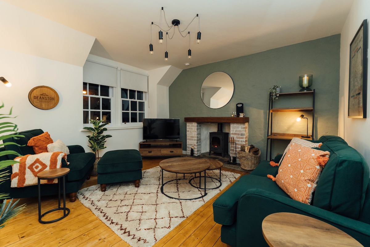 A cozy living area features a green sofa set accompanied by a decorative ottoman. A round coffee table sits on a textured rug, with a circular mirror enhancing the space. A modern TV is positioned near a fireplace, providing a warm focal point.
