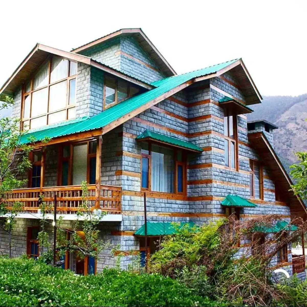 A wooden chalet features a combination of stone and wood exteriors, highlighted by green roofing. Large windows allow natural light to flood the interiors, while surrounding greenery enhances the serene environment. The structure blends harmoniously with the picturesque landscape.