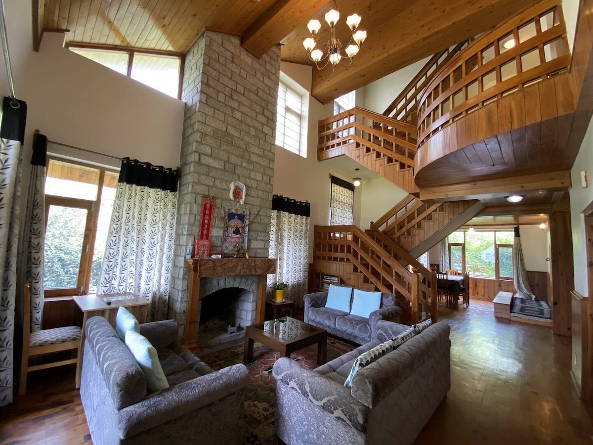 A spacious living area features high ceilings with wooden beams, showcasing large windows that allow natural light to fill the room. Two comfortable seating arrangements are centered around a stone fireplace, while an elegant staircase leads to the upper level, enhancing the open layout.